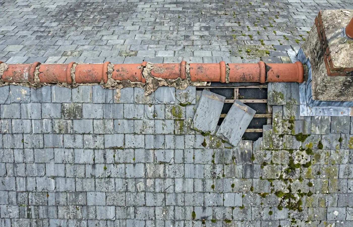 Close aerial detail of a slate roof ridge with washed out mortar joints, slipped slates below it exposing the battens and underlay, moss across the laps and a stone chimney stack with lead flashing beside