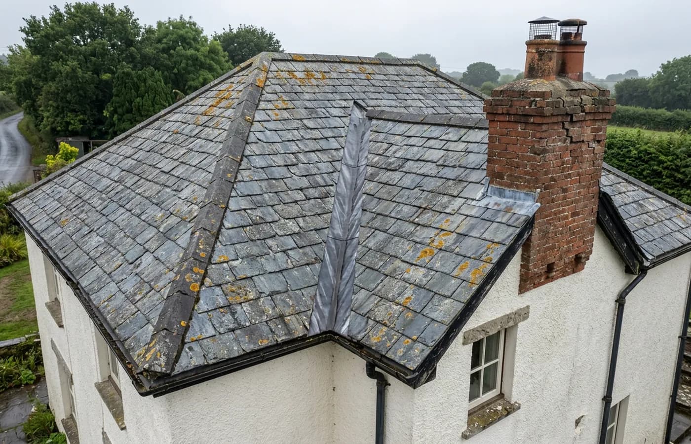 Aerial view of a rendered Devon house at a three quarter angle showing lichen spotted slates, a lead lined valley, a brick chimney stack with a cowl and black gutters